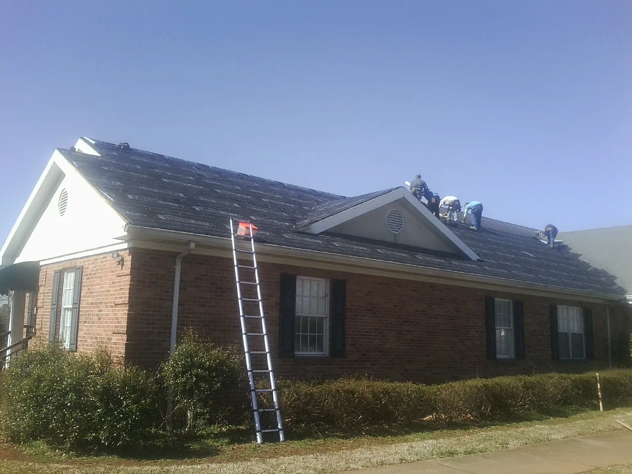 Crew tearing shingles off a brick ranch roof with a ladder set — Emergency Roof Repair work by our crew in Newton Lower Falls