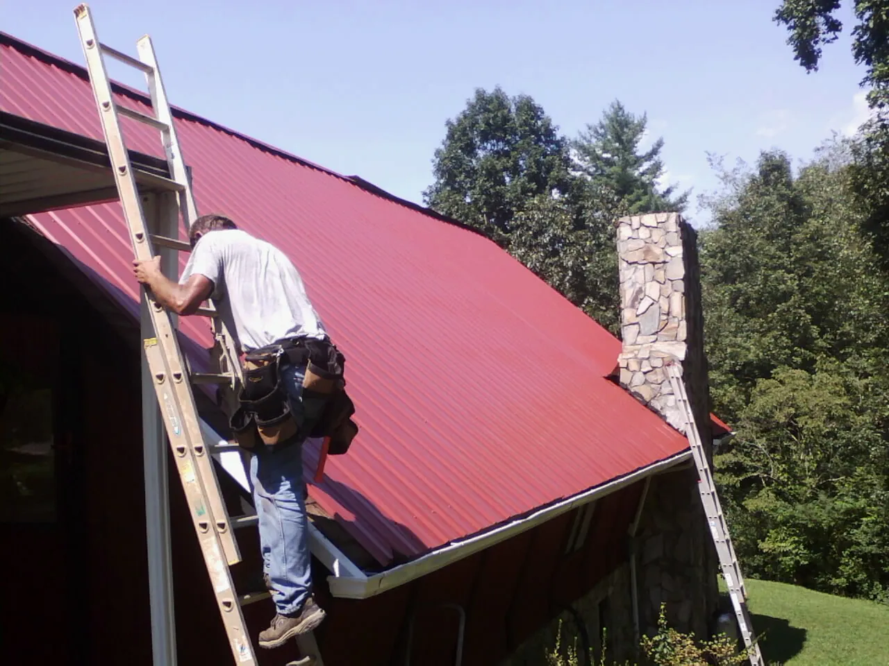 Roofer on a ladder beside a red metal roof and stone chimney — Soffit & Fascia Repair by Roofing Brothers in Newton Lower Falls
