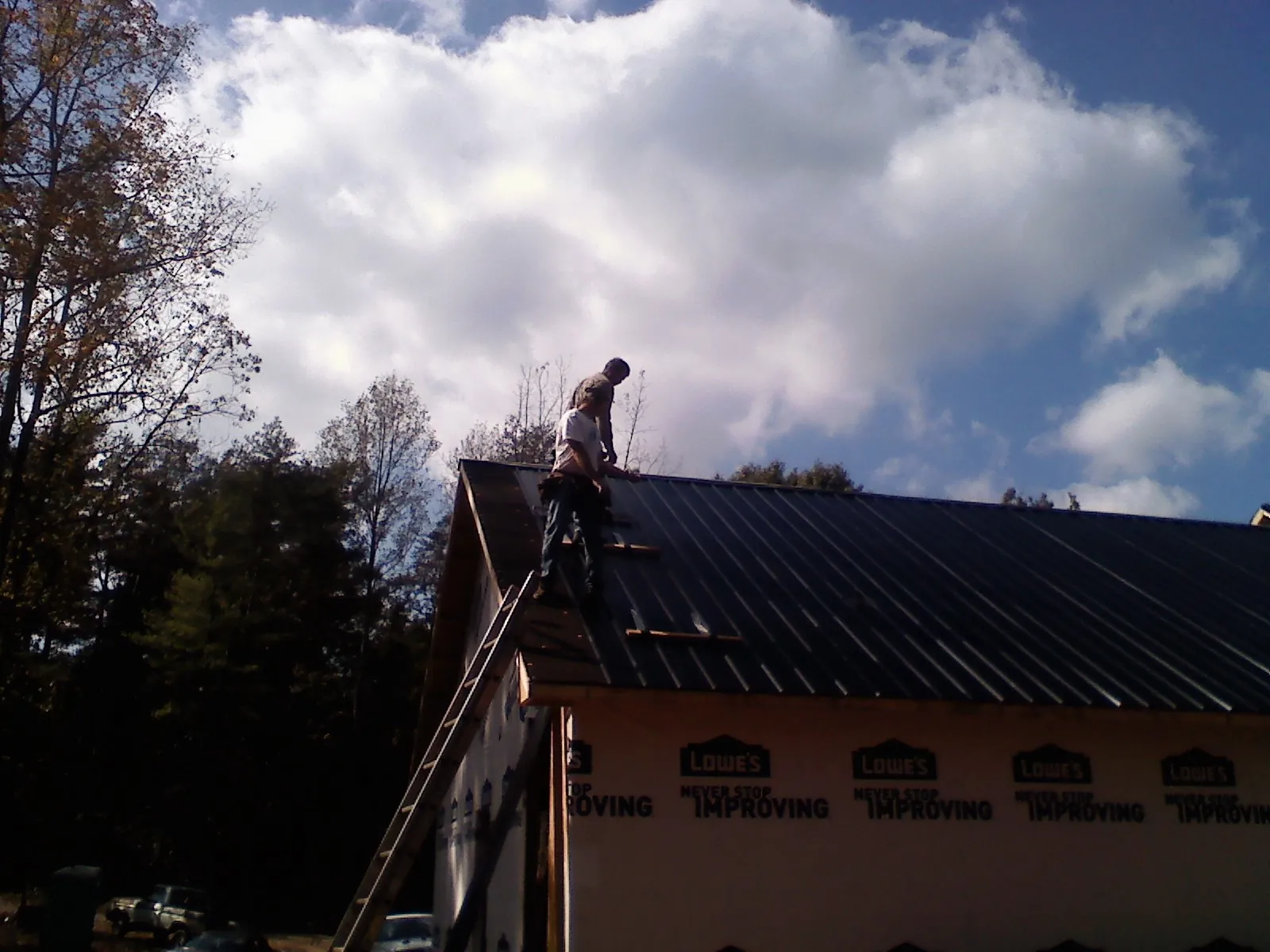 Roofer fastening dark metal panels high against a cloudy sky, from a Roof Inspection job in Newton Lower Falls